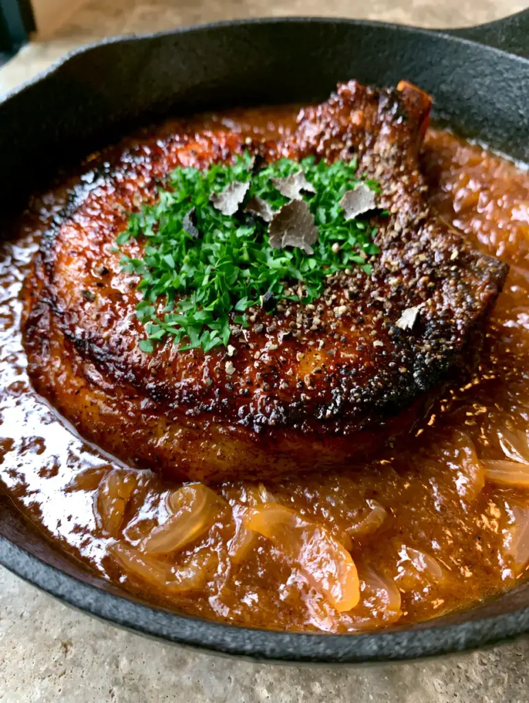 Overhead view of a smothered pork chop with translucent onion slivers in a matte black skillet.