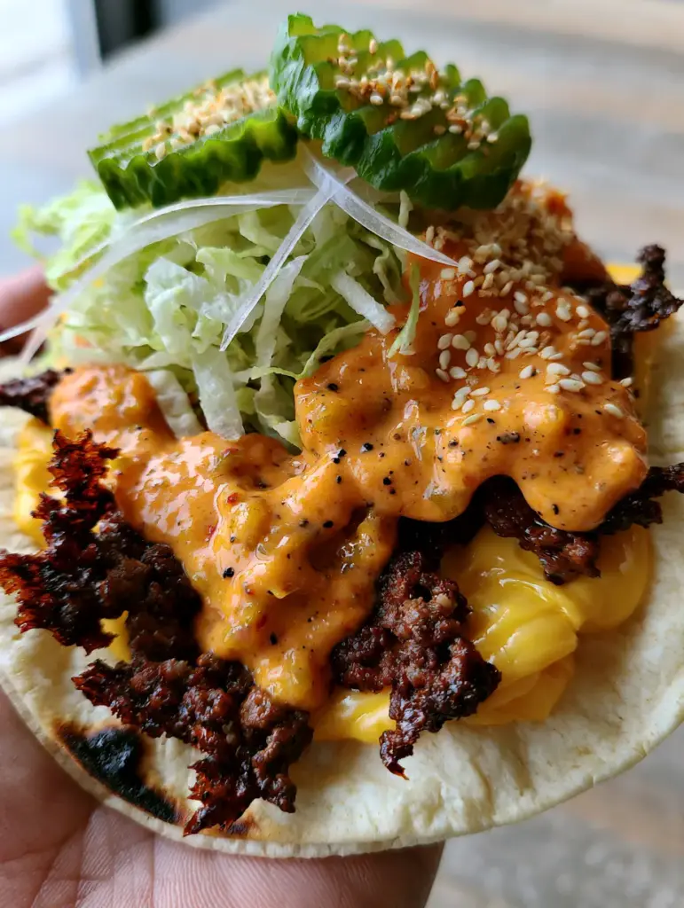A finished Smash Taco garnished with shredded iceberg lettuce, translucent white onions, crinkle-cut pickles, and toasted sesame seeds.