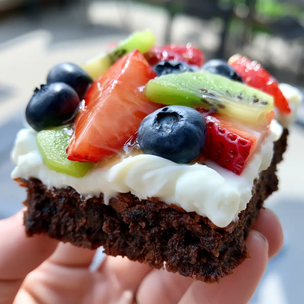 A handheld smartphone photo of a thick slice of brownie fruit pizza held up to the camera showing dense moist crumbs. (brownie fruit pizza)