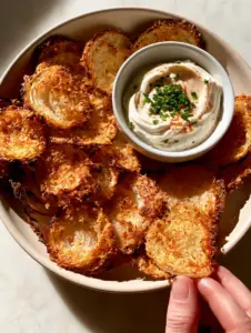 Top-down view of golden-brown crispy onion ring chips arranged in a matte bowl with a white ramekin of chive yogurt dip.