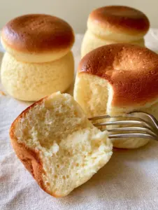 A handheld smartphone photo of three Japanese cotton cheesecake cupcakes on a light linen surface, featuring a torn cupcake revealing a micro-bubble interior.